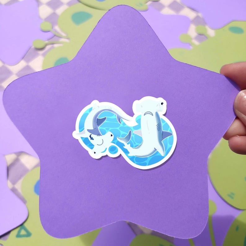 May include: A purple star-shaped sticker with a cartoon illustration of two hammerhead sharks swimming in blue water. The sharks are white with gray details. The sticker is held by a hand against a background of green and purple shapes.