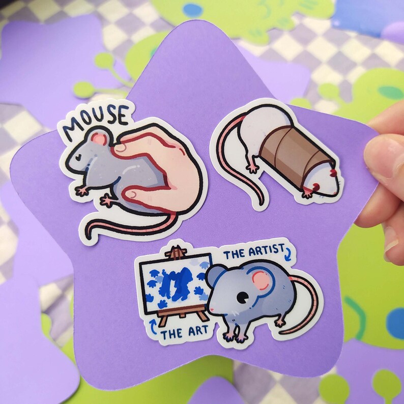 May include: A collection of mouse-themed stickers on a purple star-shaped background. The stickers feature cartoon mice in various poses, including one held in a hand, one wrapped in a brown tube, and one with an easel. Text includes "MOUSE", "THE ART", and "THE ARTIST".