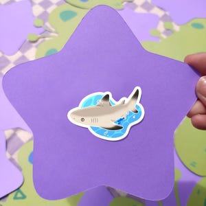 May include: A shark sticker with a light gray body, black fins, and white accents is centered on a purple star-shaped cutout. The sticker is surrounded by a blue water design. The background features green and purple shapes.