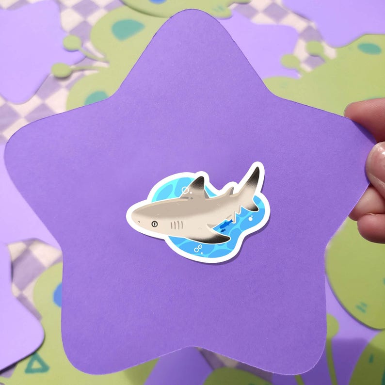 May include: A shark sticker with a light gray body, black fins, and white accents is centered on a purple star-shaped cutout. The sticker is surrounded by a blue water design. The background features green and purple shapes.