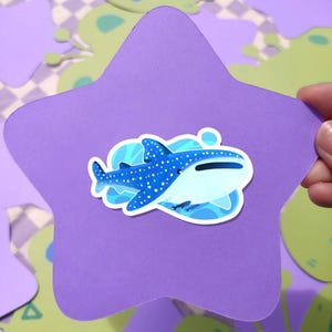 May include: A blue and white whale shark sticker on a purple star-shaped background. The sticker features a detailed illustration of the whale shark with white spots and a light blue ocean design. The background has a playful, colorful design.