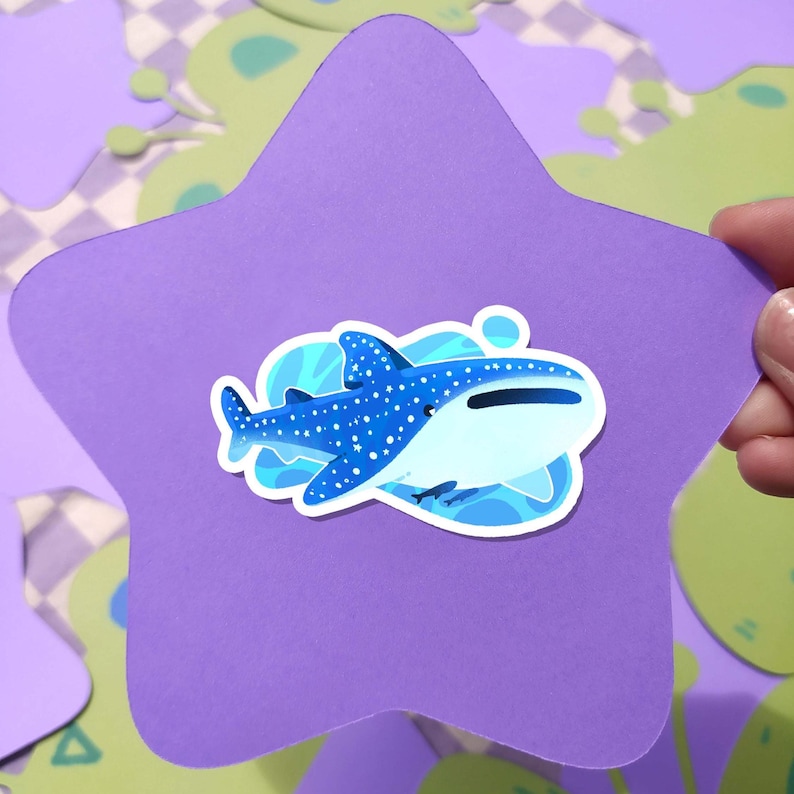 May include: A blue and white whale shark sticker on a purple star-shaped background. The sticker features a detailed illustration of the whale shark with white spots and a light blue ocean design. The background has a playful, colorful design.