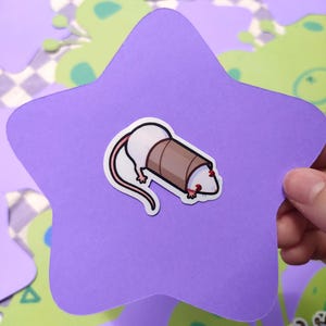 May include: A sticker featuring a white rat with a brown tube over its head, set against a purple star-shaped background. The rat has red eyes and a long, curved tail. The background includes green and white decorative shapes.