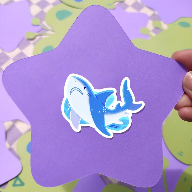 May include: A blue and white shark sticker on a purple star-shaped cutout. The shark has a detailed design with a white belly and a blue outline. The background features green and purple shapes.