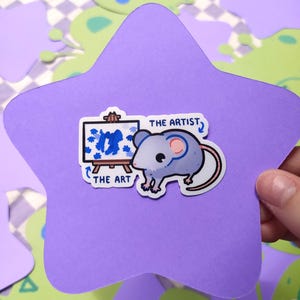 May include: A sticker featuring a cartoon mouse with a paintbrush and easel, with the text "THE ARTIST, THE ART". The sticker is on a purple star-shaped background, with green and white shapes in the background.