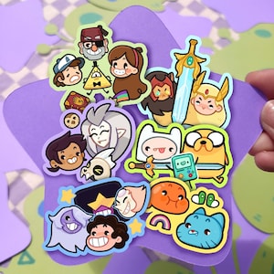 May include: A collection of colorful cartoon character stickers, including characters from Adventure Time, Steven Universe, and Gravity Falls, arranged on a purple star-shaped card. The stickers feature various shapes and sizes, with vibrant colors and detailed designs.
