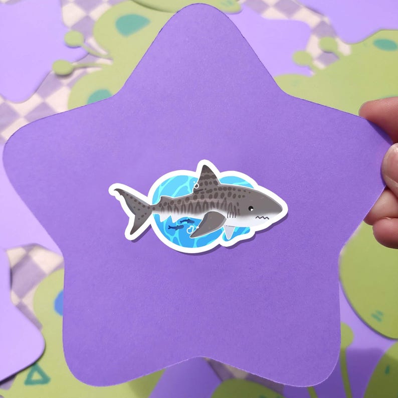 May include: A purple star-shaped sticker featuring a cartoon tiger shark design. The shark is gray with dark spots and is set against a blue background with small fish. The sticker is held against a background with green and purple shapes.