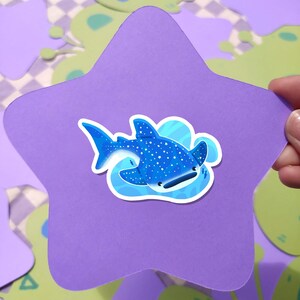 May include: A blue and white whale shark sticker on a purple star-shaped background. The sticker features a cartoon whale shark with white spots and is surrounded by blue water. The background has green and white shapes.
