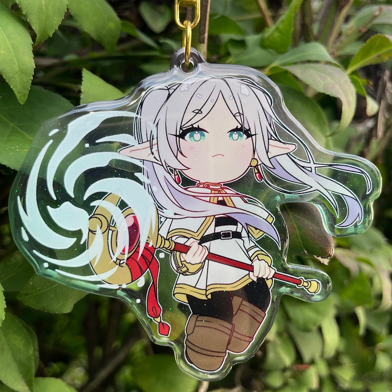 May include: A clear acrylic keychain featuring a chibi anime character with white hair, teal eyes, and elf ears. The character wears a white and gold outfit and holds a red and gold staff. The keychain has a gold-colored chain.