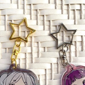 May include: Two star-shaped keychains. One is gold-colored, the other is silver. Both have a clasp and a small chain. The keychains are attached to illustrated charms with anime-style characters. The background is a white woven pattern.