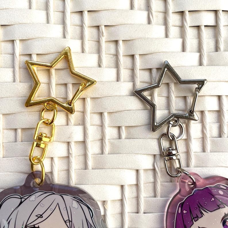 May include: Two star-shaped keychains. One is gold-colored, the other is silver. Both have a clasp and a small chain. The keychains are attached to illustrated charms with anime-style characters. The background is a white woven pattern.