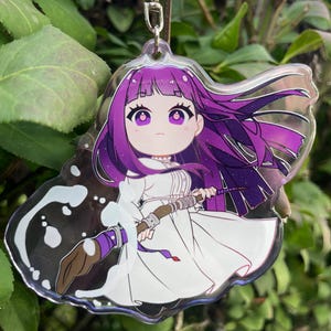 May include: A clear acrylic charm featuring a cartoon character with purple hair and eyes, wearing a white dress. The character holds a paintbrush with purple and brown accents. The charm has a silver keychain attachment and is set against a green background.