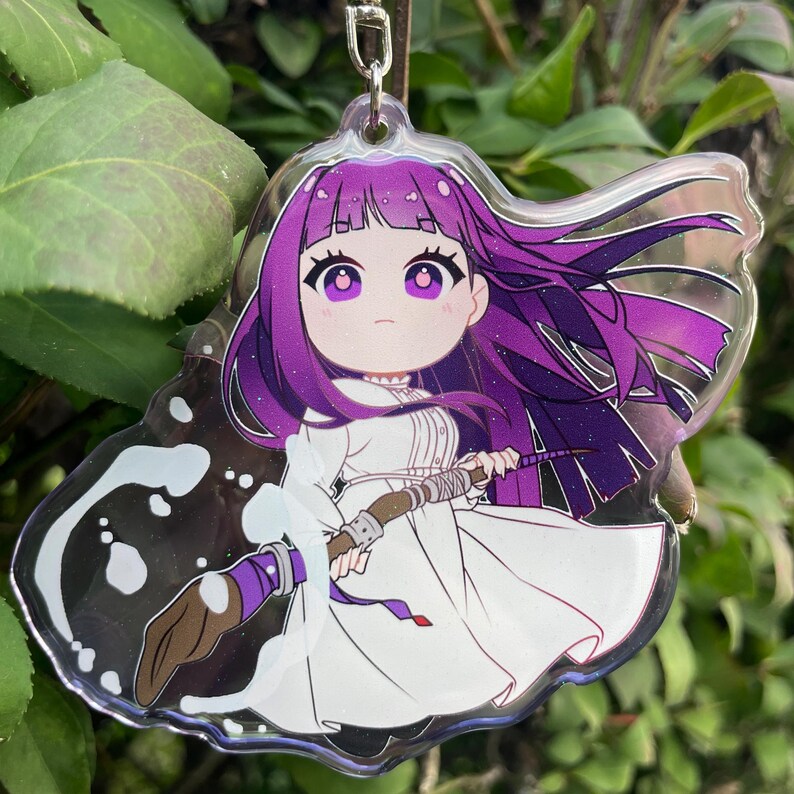 May include: A clear acrylic charm featuring a cartoon character with purple hair and eyes, wearing a white dress. The character holds a paintbrush with purple and brown accents. The charm has a silver keychain attachment and is set against a green background.