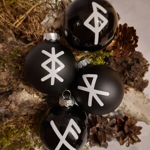 Könnte beinhalten: Vier schwarze Weihnachtskugeln mit weißen Runen darauf. Die Kugeln sind auf einem Bett aus Moos und Tannenzapfen angeordnet.