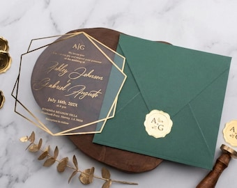 Invitación de boda acrílica con sobre verde esmeralda y lámina dorada, diseño geométrico transparente, sello de monograma, papelería moderna de lujo.