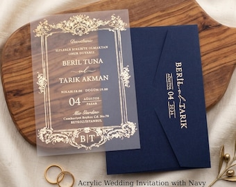 Invitación de boda acrílica con sobre azul marino y detalles en lámina dorada. Escudo con monograma dorado. Tarjeta de boda transparente para una boda clásica.
