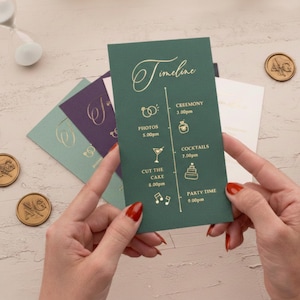 May include: A dark green wedding timeline card with gold lettering and icons, held by a person with red nail polish. The card lists event times for photos, ceremony, cocktails, cake cutting, and party time. Other cards and wax seals are in the background.