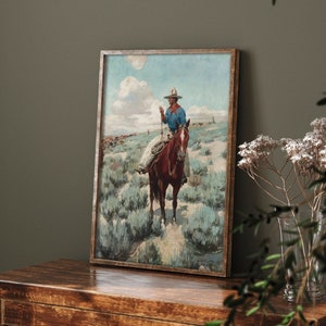 May include: A vintage framed painting of a cowboy riding a brown horse through a field of sagebrush. The cowboy is wearing a blue shirt, a brown hat, and a red bandana. The painting is set against a blue sky with white clouds.