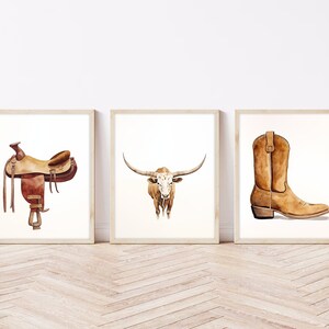 May include: Three watercolour paintings in wooden frames. The first painting shows a brown leather saddle. The second painting shows a white and brown longhorn steer. The third painting shows a brown cowboy boot.