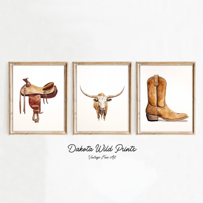 May include: Three watercolour paintings in gold frames. The first painting shows a brown leather saddle. The second painting shows a brown and white longhorn steer. The third painting shows a brown cowboy boot. The text "Dakota Wild Prints" and "Vintage Fine Art" is below the paintings.