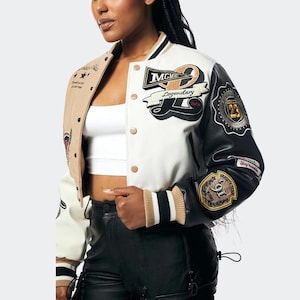 Könnte beinhalten: Eine Collegejacke mit weißem Rumpf, beigem rechtem Ärmel und schwarzem linkem Ärmel. Sie ist mit gestickten Aufnähern versehen, die Texte wie MCMXC, Legendary und 23 zeigen. Die Jacke hat eine Druckknopfleiste, gestreifte Rippbündchen und ein weißes bauchfreies Top.