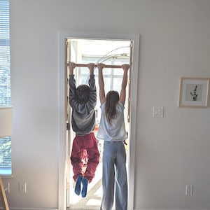 May include: Two children hanging upside down from a wooden bar installed in a doorway. One child wears a gray hoodie and red pants, the other wears a light blue shirt and gray pants. The room has white walls and a wooden floor.