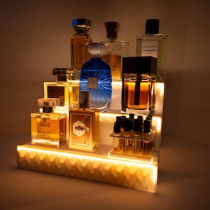 Op de afbeelding: Een wit, meerlagig displayrek met verschillende glazen parfumflesjes. De flesjes zijn gevuld met verschillende gekleurde vloeistoffen en hebben verschillende etiketten. Het rek is verlicht met warmwitte LED-verlichting.