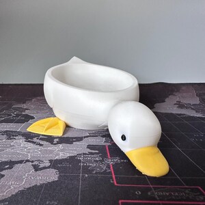 Puede incluir: Un recipiente blanco con forma de pato con pico y patas amarillas. El recipiente tiene forma de cuenco y la cabeza del pato est&aacute; separada del cuerpo. El pato est&aacute; hecho de un material liso y s&oacute;lido.
