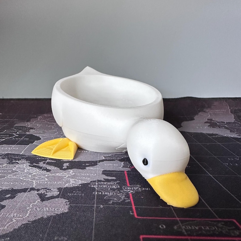 Puede incluir: Un recipiente blanco con forma de pato con pico y patas amarillas. El recipiente tiene forma de cuenco y la cabeza del pato est&aacute; separada del cuerpo. El pato est&aacute; hecho de un material liso y s&oacute;lido.