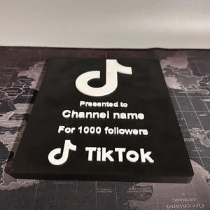 Peut inclure: Une plaque noire avec un logo TikTok blanc et le texte "Pr&eacute;sent&eacute; &agrave; Channel name Pour 1000 abonn&eacute;s TikTok".