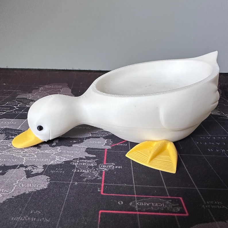 Puede incluir: Un recipiente blanco con forma de pato con pico amarillo y una peque&ntilde;a pata amarilla. El recipiente est&aacute; dise&ntilde;ado para contener objetos. El pato est&aacute; sobre un fondo de mapa del mundo.