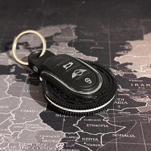 May include: Black car key chain with a silver Mini Cooper logo and a tyre tread design. The keychain is on a black and grey map.