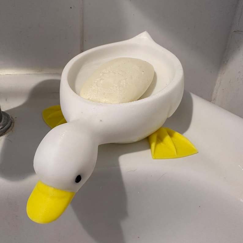 Puede incluir: Un jabonera blanca con forma de pato con pico y patas amarillas. La jabonera contiene una pastilla de jab&oacute;n. La jabonera est&aacute; hecha de cer&aacute;mica o pl&aacute;stico y est&aacute; dise&ntilde;ada para colocarse en un lavabo de ba&ntilde;o.