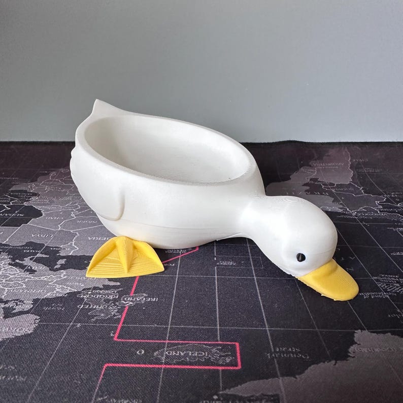 Puede incluir: Una maceta blanca con forma de pato con pico amarillo y un peque&ntilde;o accesorio amarillo. La maceta tiene un cuerpo grande en forma de cuenco y un cuello curvado hacia abajo. El ojo del pato es un peque&ntilde;o punto negro.