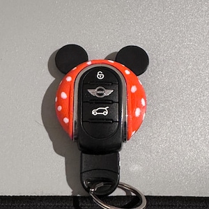 May include: A black Mini Cooper car key with a red cover featuring white polka dots. The key has a silver key ring and is adorned with black, rounded ears, creating a whimsical design.