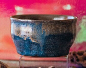 May include: A blue and brown ceramic bowl with a glossy finish sits on a clear acrylic stand. The bowl is surrounded by seashells on a sandy surface.