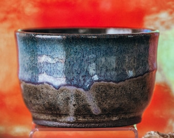 May include: A handcrafted ceramic bowl with a dark blue and brown glaze. The bowl has a wide, slightly flared shape and sits on a clear base. The background is a gradient of orange and red.