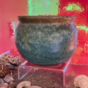 May include: A teal green ceramic pot with a brown rim, sitting on a clear acrylic stand. The pot has a speckled, textured surface and is displayed on a sandy surface with seashells.