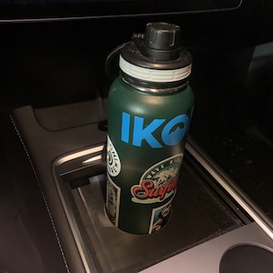 May include: A green and blue water bottle with the word "IKO" printed on it. The bottle has a black lid and a white band around the middle. The bottle is sitting in a cup holder in a car.