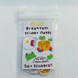 May include: A clear plastic bag containing a sheet of breakfast-themed stickers. The stickers feature illustrations of pancakes, eggs, and frogs. Text on the sheet reads "Breakfast sticker flakes" and "I'm still really sleepy?" with "20+ Stickers!!".