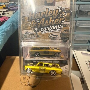 May include: A gold-colored '64 Chevy Nova Wagon Gasser diecast toy car in a clear plastic blister pack. The packaging features the text 'Harley & Asher customs' and 'EST. 2019 - PREMIUM DIECAST MODS'. The car has 'Gold Prospector' printed on the side.