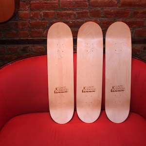 May include: Three blank wooden skateboards with the text "Urban Outfitters" printed on the bottom of each board. The skateboards are sitting on a red couch.