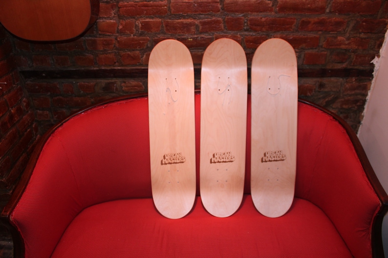 May include: Three blank wooden skateboards with the text "Urban Outfitters" printed on the bottom of each board. The skateboards are sitting on a red couch.