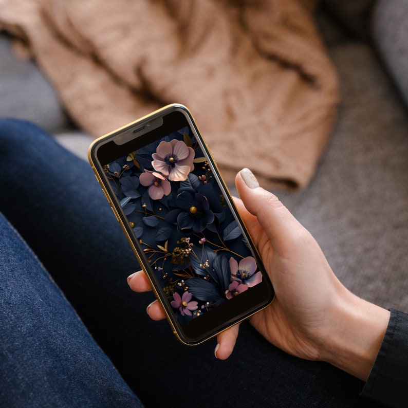 May include: A smartphone displaying a floral wallpaper with pink and white flowers on a dark blue background. The flowers are surrounded by dark green leaves and gold accents.