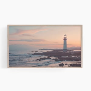 Puede incluir: Un faro blanco se alza sobre una costa rocosa, iluminado por el cálido resplandor del sol poniente. El cielo es una suave mezcla de rosa y naranja, reflejándose en las tranquilas olas del océano.