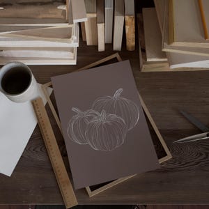 May include: A framed print with a minimalist line drawing of three pumpkins on a brown background.