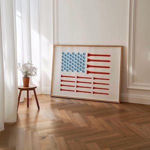 Puede incluir: Obra de arte enmarcada que representa la bandera estadounidense, compuesta por bolas de golf azules y tees de golf rojos. La obra de arte se exhibe en un marco de madera clara, apoyada contra una pared blanca. La habitación tiene un suelo de madera y una mesita auxiliar.