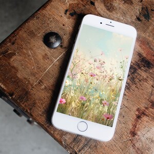 May include: A white smartphone with a floral wallpaper. The wallpaper features a field of pink and white flowers with a blue sky in the background.