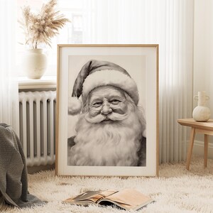 May include: A framed black and white portrait of Santa Claus. The image features a detailed depiction of Santa's face, with a long white beard, round glasses, and a traditional hat. The artwork is displayed in a light-colored wooden frame, suitable for holiday decor.