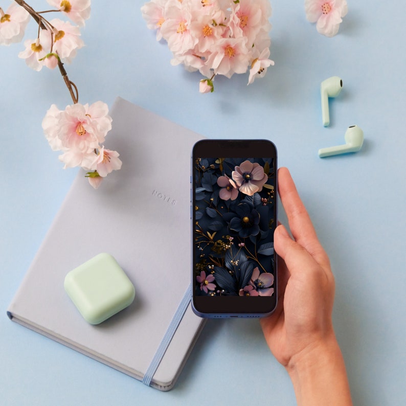 May include: A blue smartphone with a floral wallpaper on the screen. The phone is being held by a hand. A light blue notebook with a white elastic band is in the background.